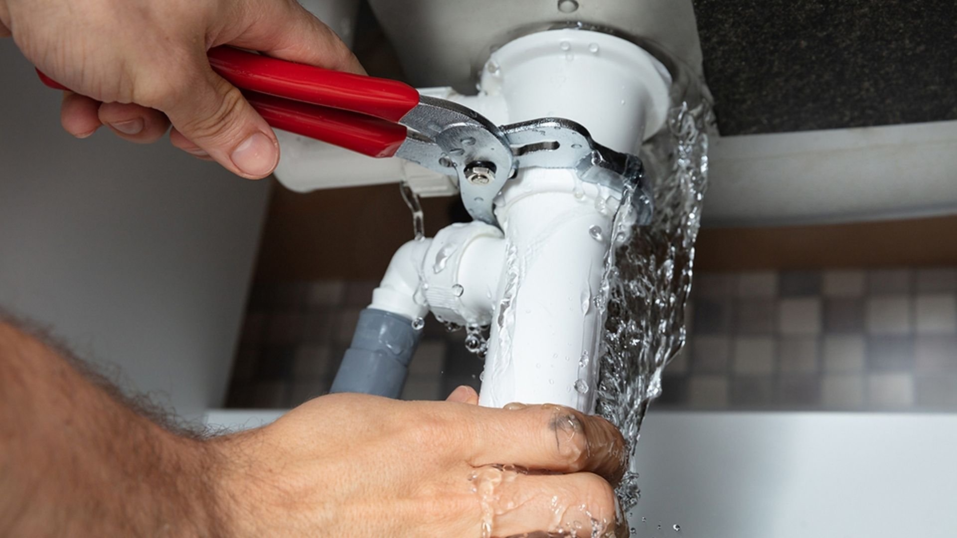 Person using red wrench to tighten white under-sink plumbing valve with water splashing