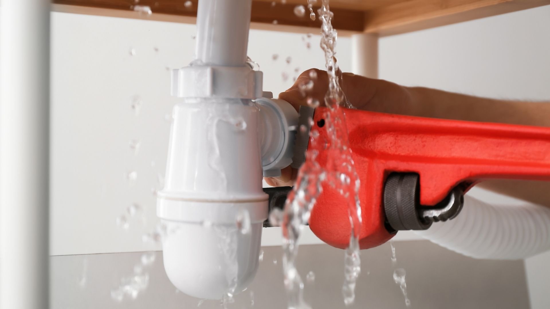 Plumber using red wrench to fix white PVC pipe connection with water spraying