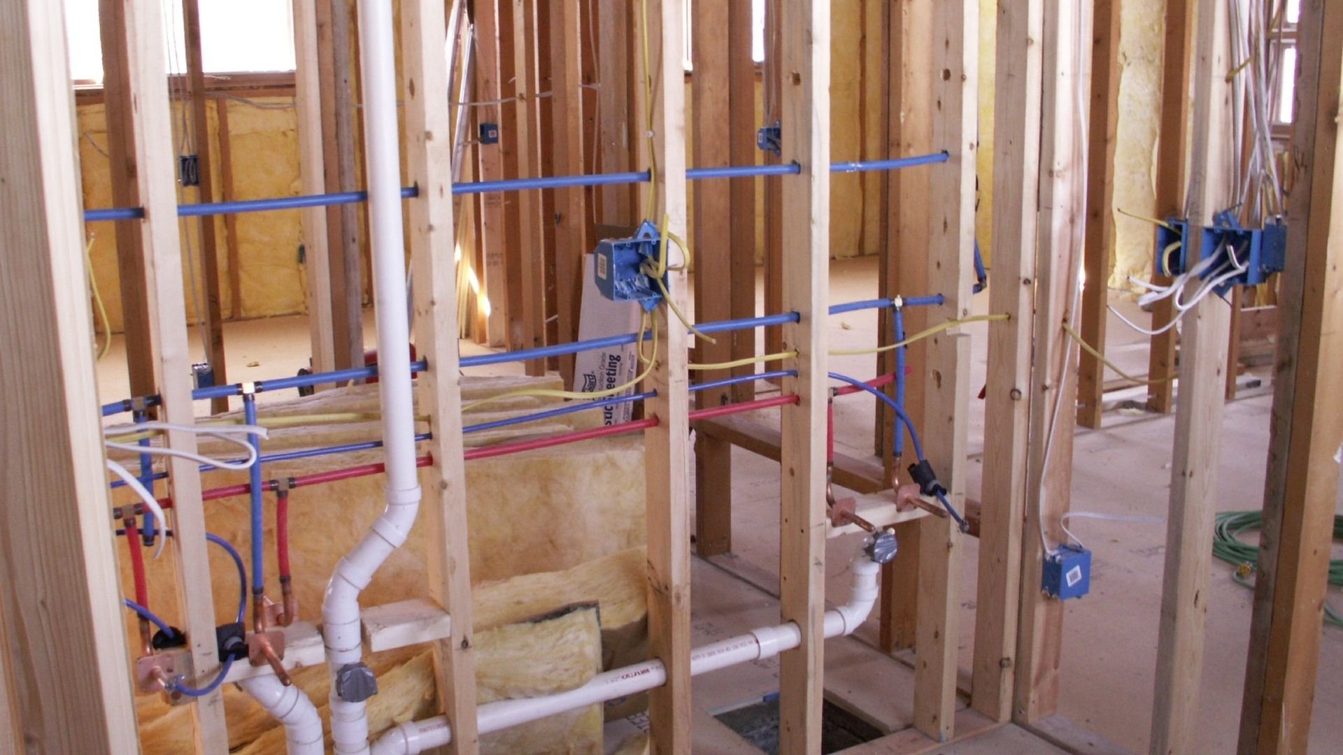 Under-construction home interior with wooden framing, blue electrical conduit, and plumbing pipes.